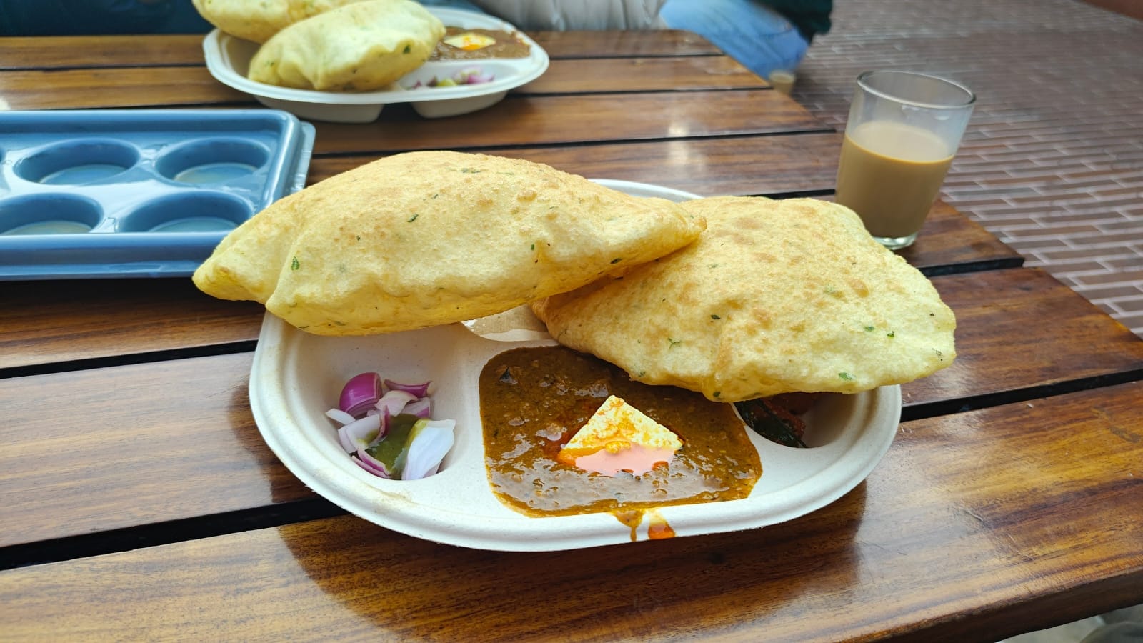 A plate of puffed bread and curry with chopped onions and a glass of tea on a wooden table.