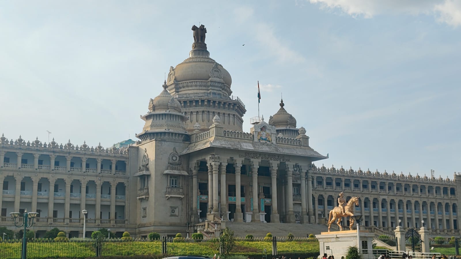 An ornate stone government building with domes, columns, an Indian flag, and a golden equestrian statue.