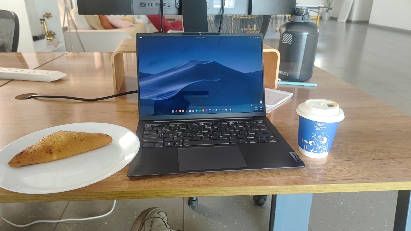 A laptop on a wooden desk beside a coffee cup and a plate with a snack.