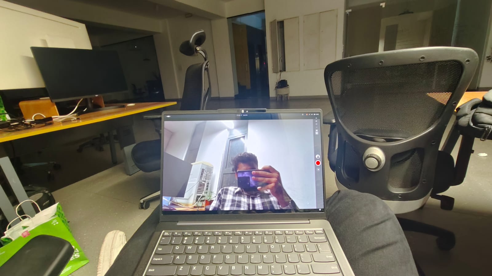 A laptop showing a camera view of someone holding a phone, surrounded by office chairs and monitors.