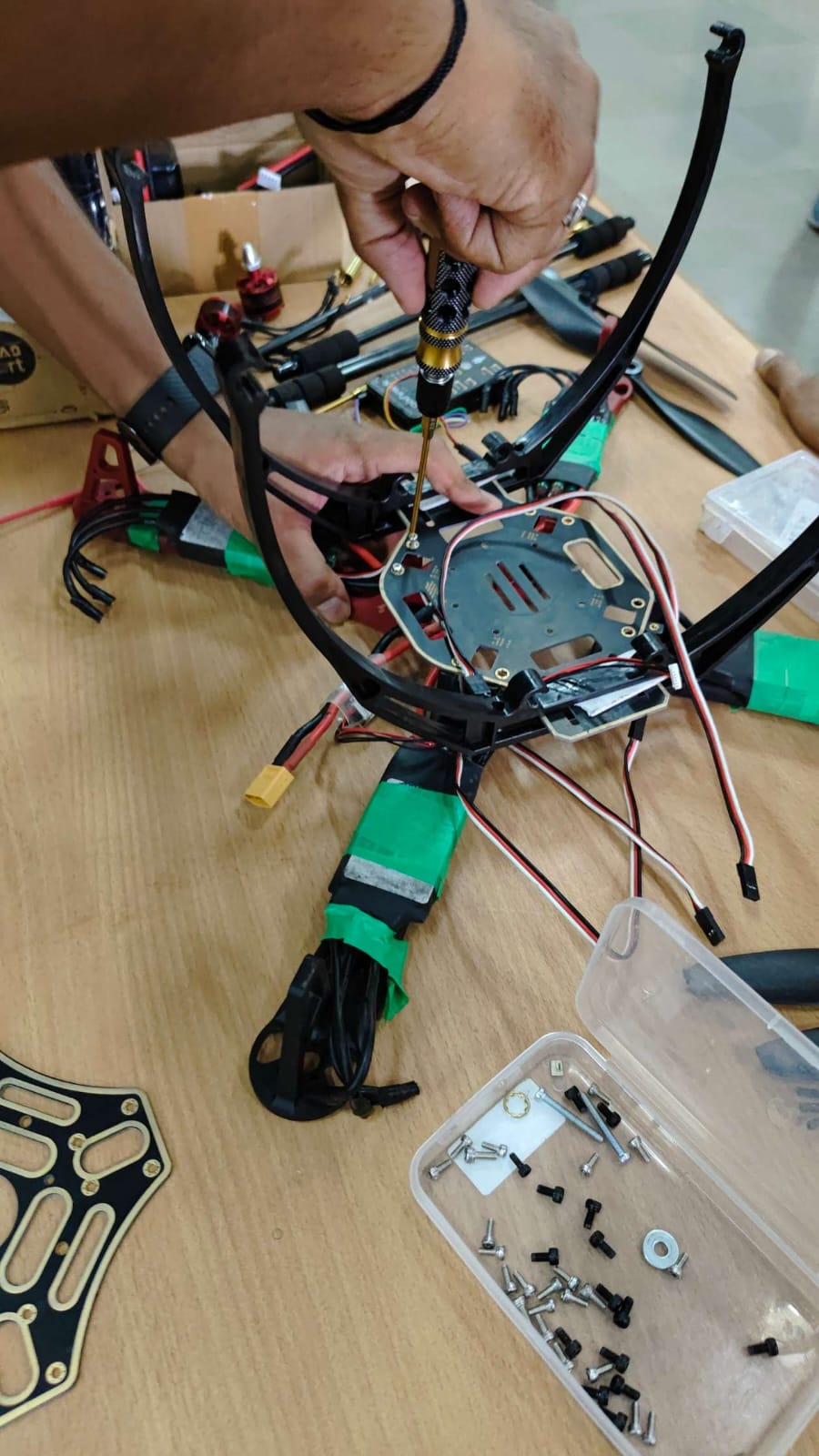 Hands using a screwdriver to assemble a drone frame, surrounded by motors, wires, batteries, and spare screws.