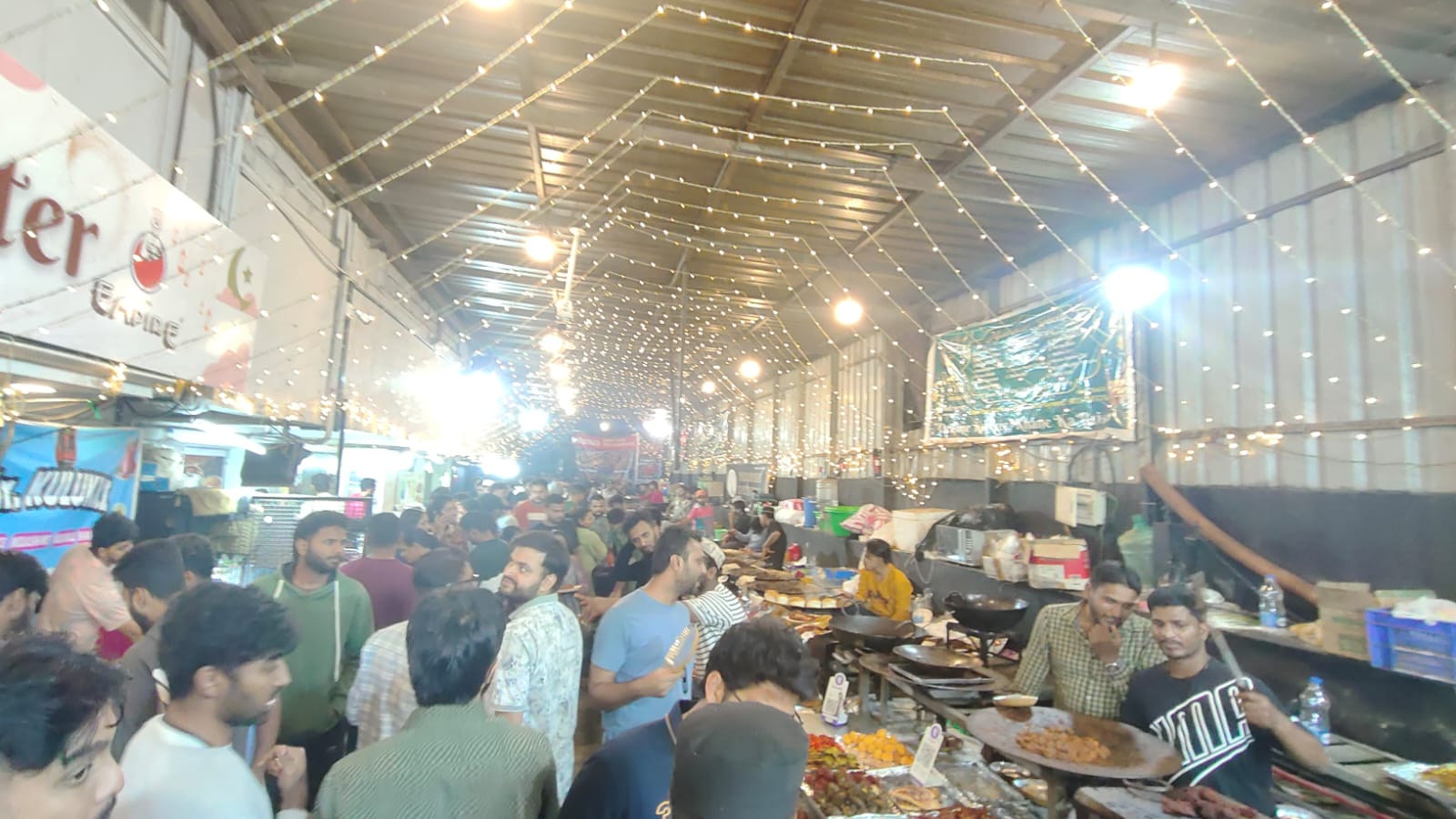 A busy indoor food market with stalls, customers, and strings of warm overhead lights.