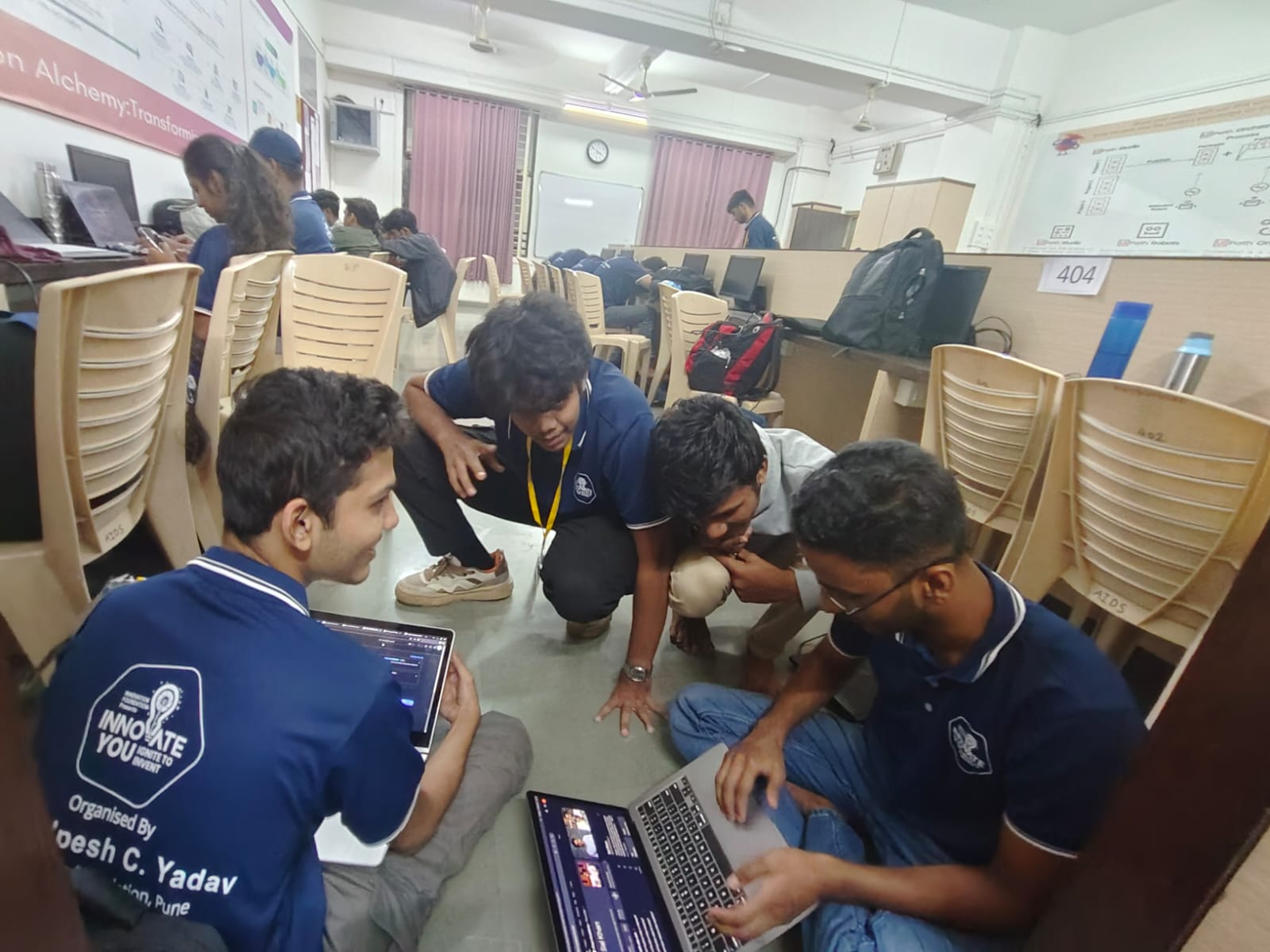 A group gathered around laptops on the floor of a computer lab, with chairs and workstations behind them.